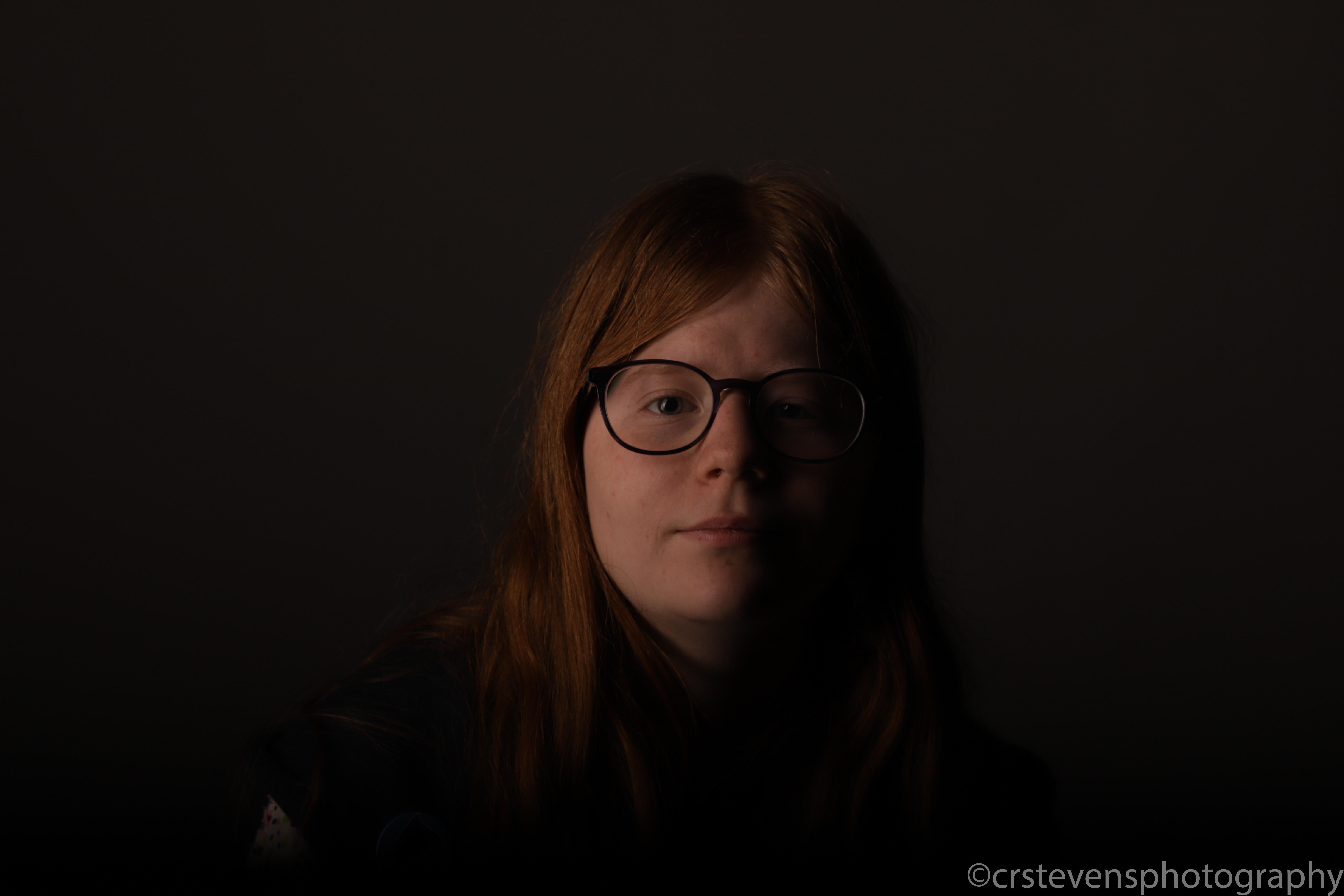 someone with glasses looking at the camera and dim lighting. only one side of their face is visible