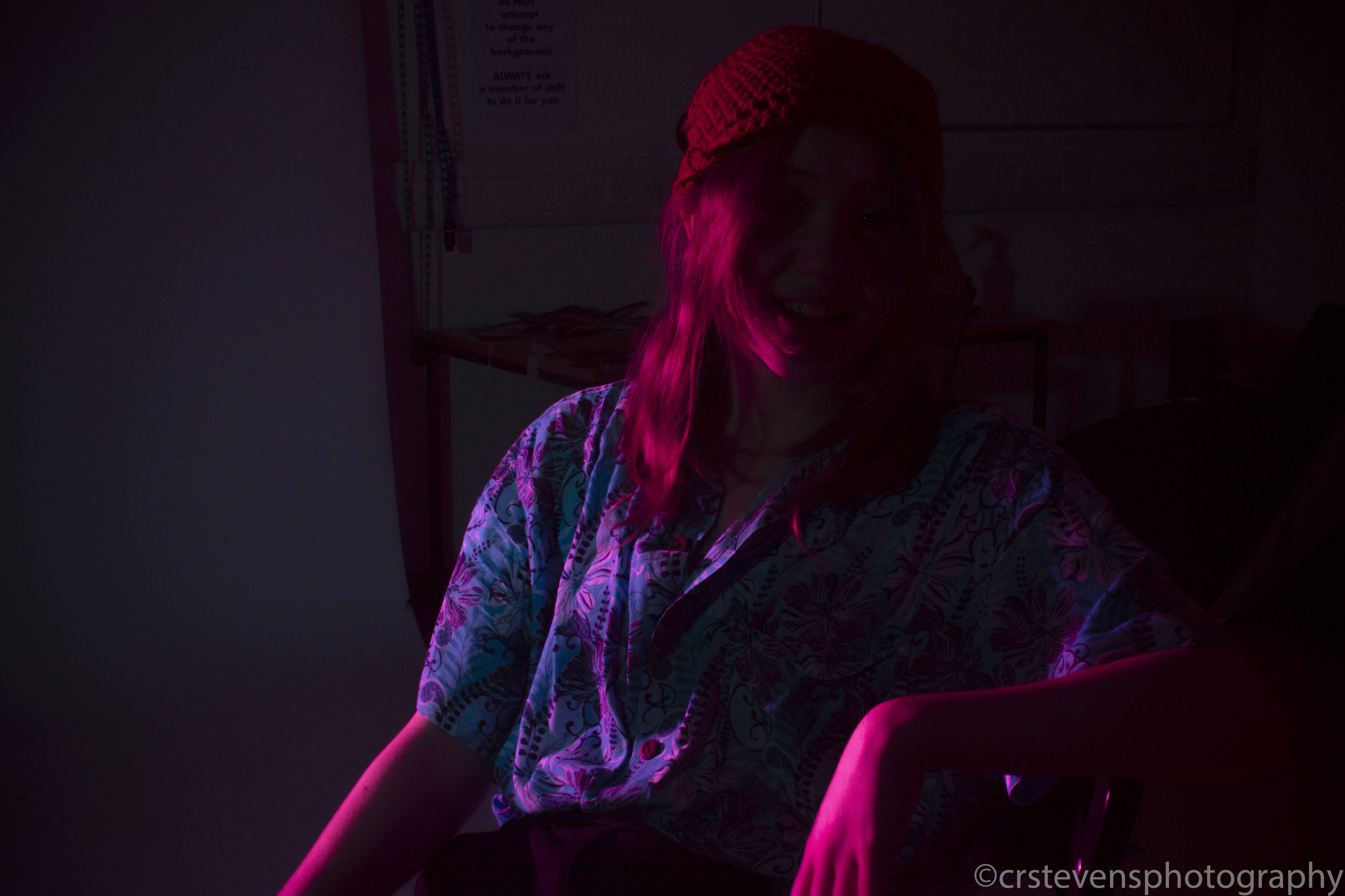 a person with a beanie and a patterned shirt in bisexual lighting leaning to one side with their face hidden
