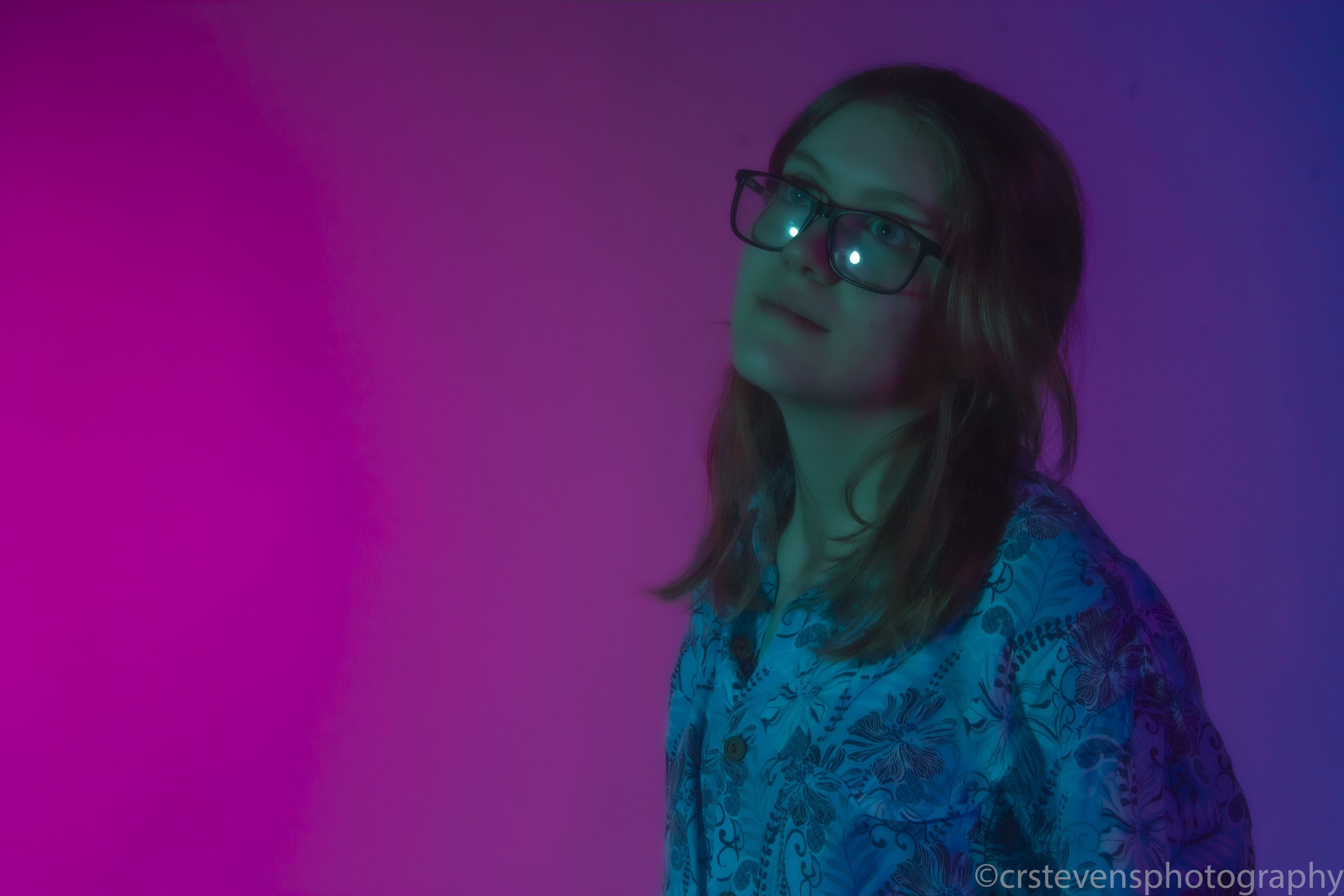 a person with a patterned shirt and glasses looking past the camera with their body turned away in bisexual lighting
