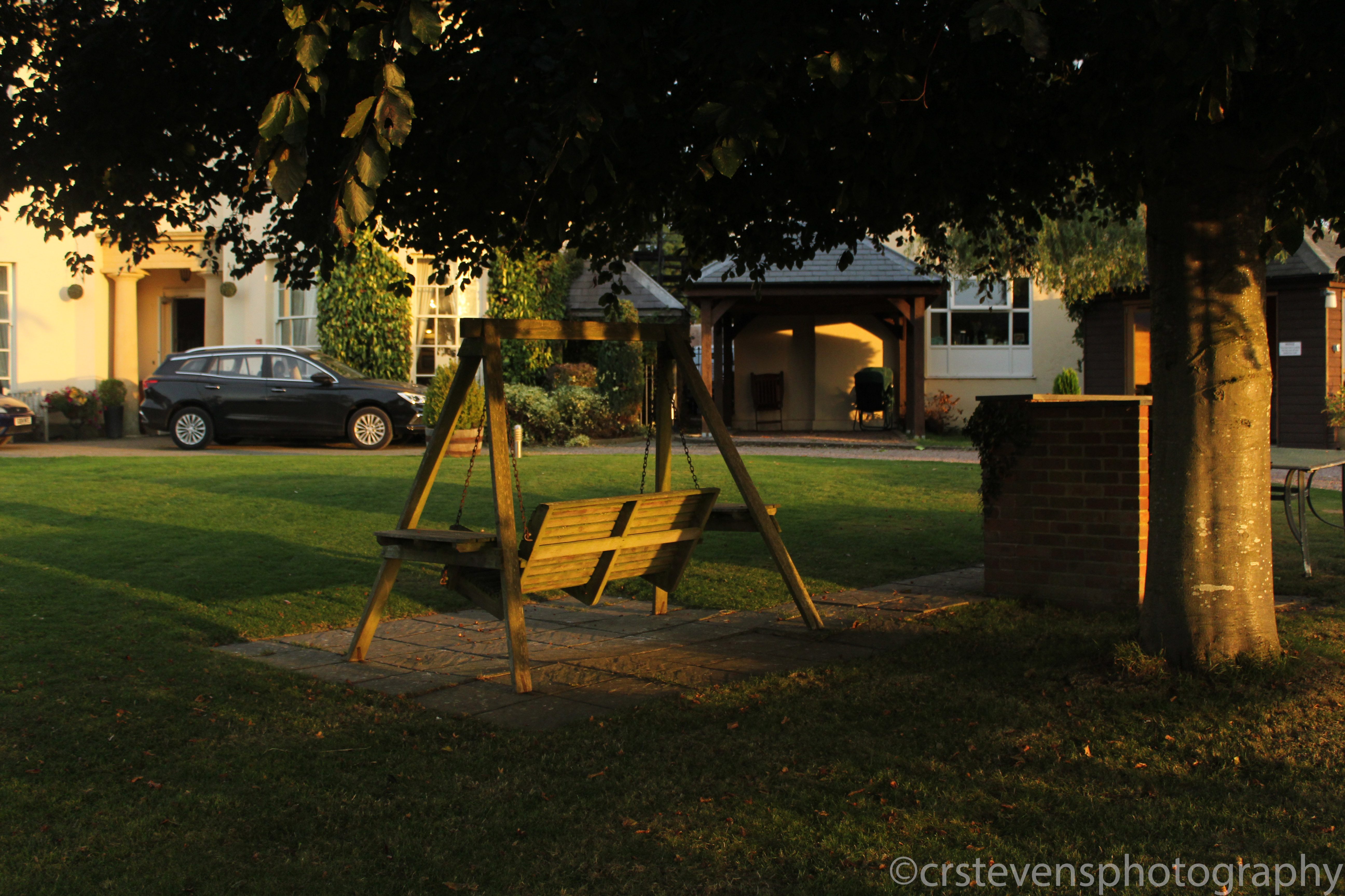 a wooden bench swing next to a tree in front of a large house and some cars at golden hour